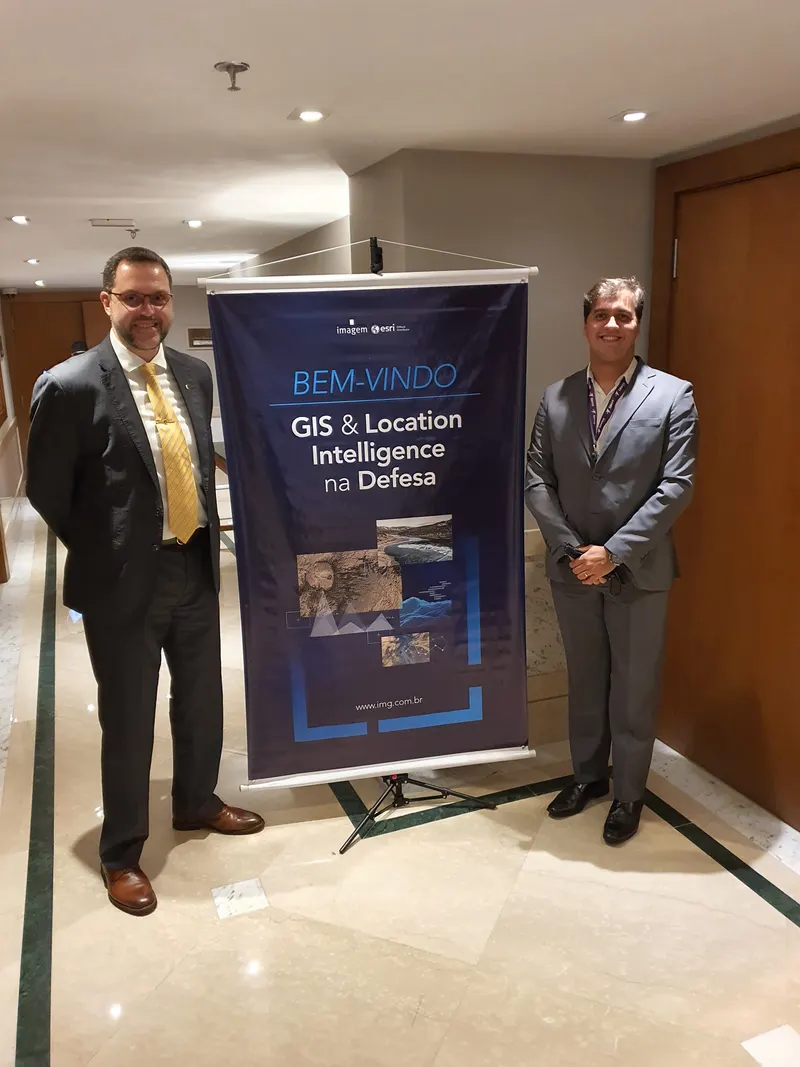 Dois participantes ao lado do banner do evento GIS e Location Intelligence na Defesa, em Brasília